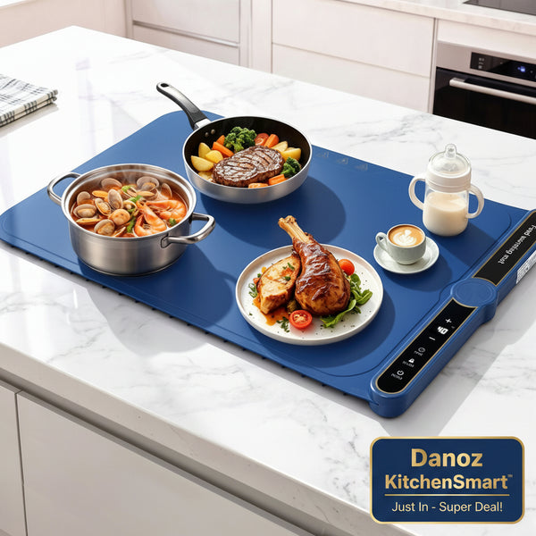 A blue electric hot plate on a white marble kitchen countertop, shown with a frying pan, a pot of seafood, a plate of grilled chicken, and a small milk bottle. The hot plate has a digital control panel on the right side.