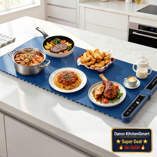 A blue electric skillet/tray sits on a white kitchen countertop, surrounded by prepared meals and a baby bottle. The scene is set in a bright, modern kitchen with white cabinets and marble surfaces.
