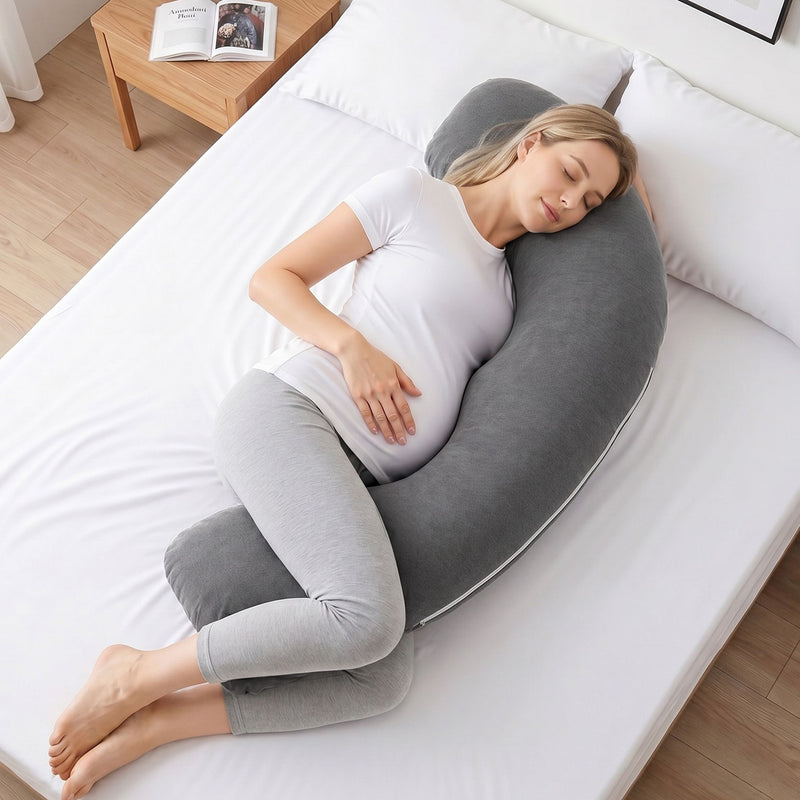 A pregnant woman sleeping soundly on a bed, supported by a dark gray U-shaped maternity pillow. She is wearing a white t-shirt and light gray leggings.