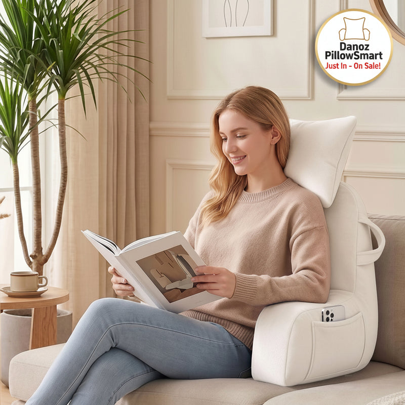 A woman sits comfortably in a white ergonomic lounge chair on a beige sofa, holding an open book. The chair has a built-in headrest, side pockets, and a phone holder, set in a bright, modern living room with a potted plant and curtains.