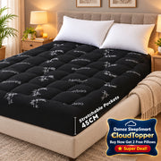 A black tufted mattress topper or mattress shown on a bed in a bedroom setting, with white pillows, a bedside lamp, and a green wall panel visible. The topper has a subtle white bamboo pattern and text reading 'Stretchable Pockets 45CM'.