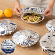 Person unwrapping a bowl of food from tinfoil on a wooden table with oranges in the background.