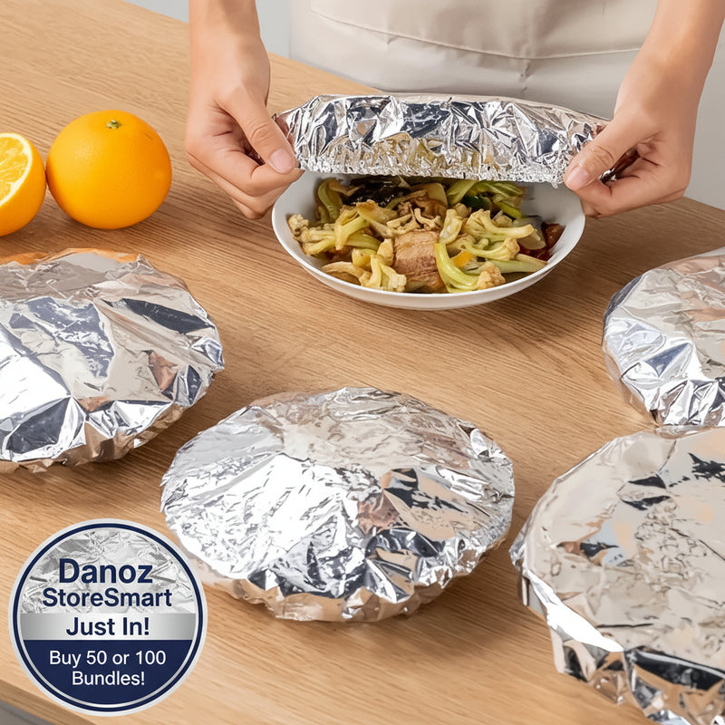 Person unwrapping a bowl of food from tinfoil on a wooden table with oranges in the background.
