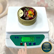 A small round tin of Tropical Fruit Drops sits on a white digital scale with a green display panel. A finger is pressing the scale's buttons, and the scale reads 79 grams.