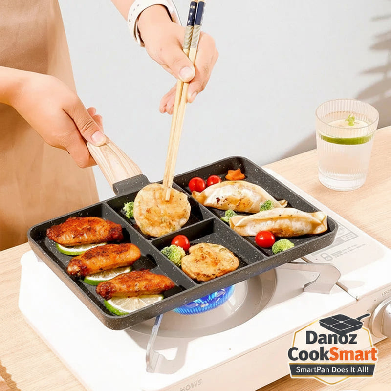 A black multi-compartment griddle pan with a wooden handle is being cooked on a portable stove. Hands are visible holding the handle and chopsticks, with food items like dumplings, pancakes, and grilled sausages cooking inside the compartments.