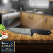 A kitchen knife with a matte gray blade and black handle resting on a wooden cutting board, with a blurred kitchen background and fresh vegetables on the left.