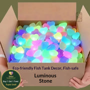A cardboard box filled with glowing, translucent heart-shaped decorative stones in multiple colors, held open by two hands against a muted olive background. Text on the box reads 'Eco-friendly Fish Tank Decor, Fish-safe Luminous Stone'.