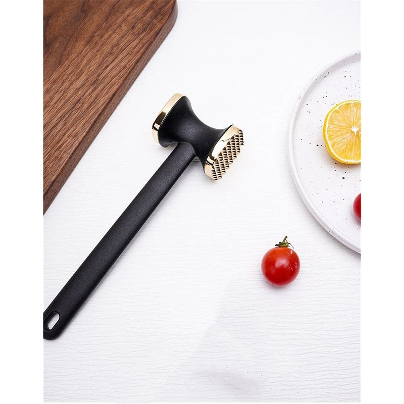 A black and gold meat mallet resting on a white textured surface, styled with a wooden cutting board, a white plate with a lemon slice, and a cherry tomato.