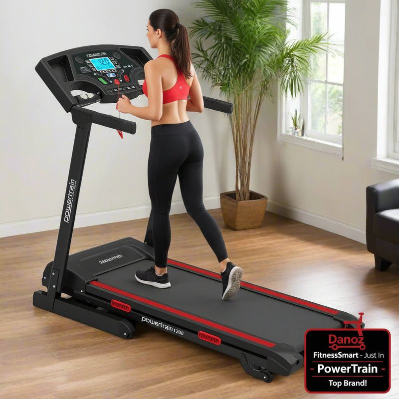A black Powertrain K200 electric treadmill with a folding design, placed in a home environment with a window and indoor plants in the background.