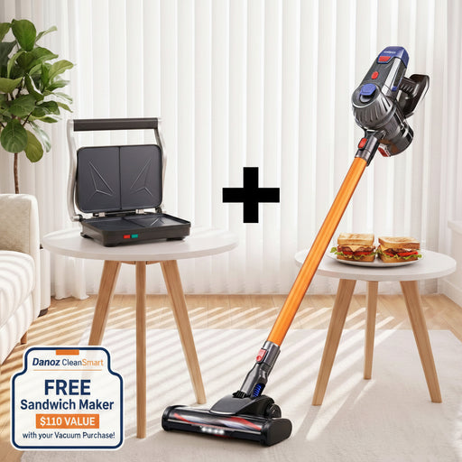 A cordless stick vacuum cleaner with an orange handle is shown on a light rug in a bright room. A small round table beside it holds an open sandwich press with two sandwiches, and a circular inset at the bottom right shows two nozzle attachments.