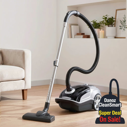 A silver and black canister vacuum cleaner with a flexible hose and floor nozzle, shown in a bright living room setting on a light wood floor next to a beige armchair.