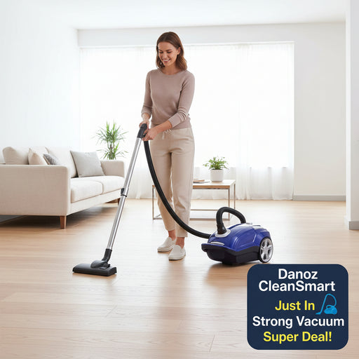 A blue and silver canister vacuum cleaner with a flexible black hose and floor nozzle, shown in a bright living room setting on a light wood floor next to a beige armchair.