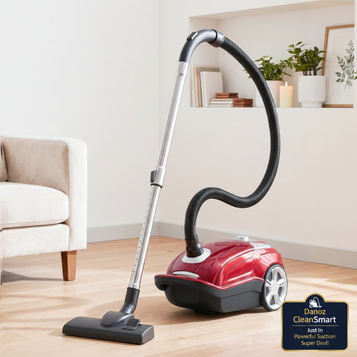 A red and black canister vacuum cleaner with a silver handle and black hose, positioned on a light wood floor in a bright, modern living room next to a beige armchair.