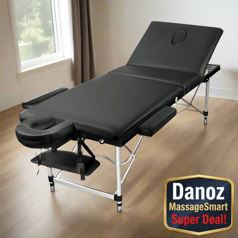 A portable 3 fold aluminium massage table in black color with a face cradle and armrest, set on a wooden floor with a window in the background.