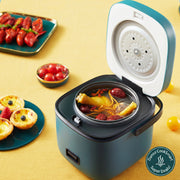 A compact blue mini rice cooker with a white non-stick pot inside, displayed on a table with some food items around it, including a steaming dish, rice spoon, and measuring cup.